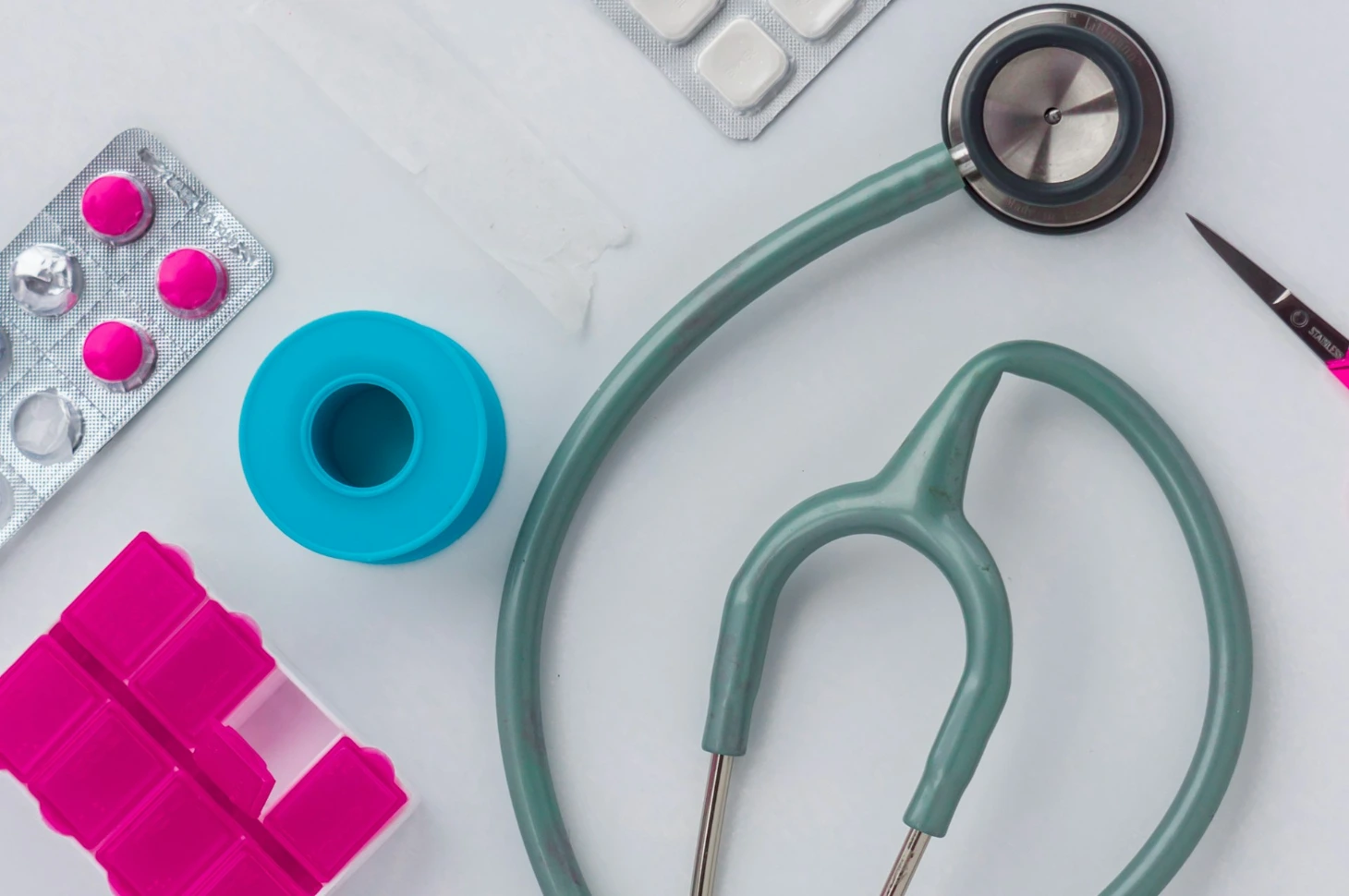 A stethoscope, pink pills, blue tape, and scissors on a white surface